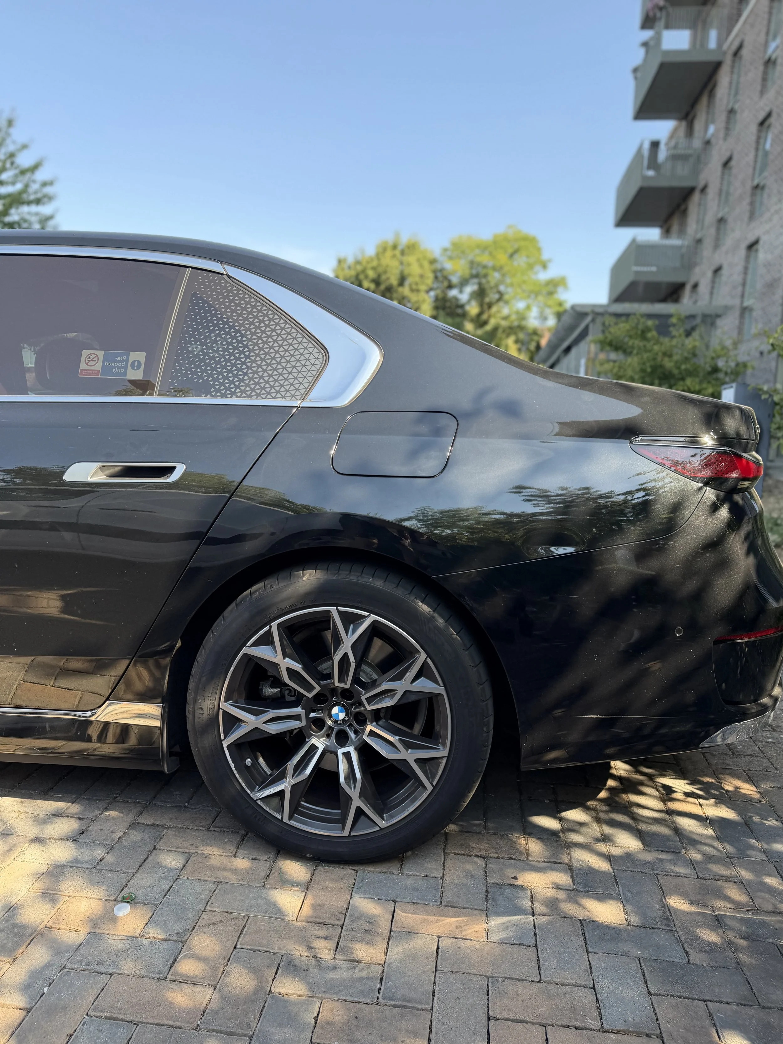 Close-up of the rear and side of a black BMW car parked on a brick driveway, with a multi-story apartment building and green trees in the background.