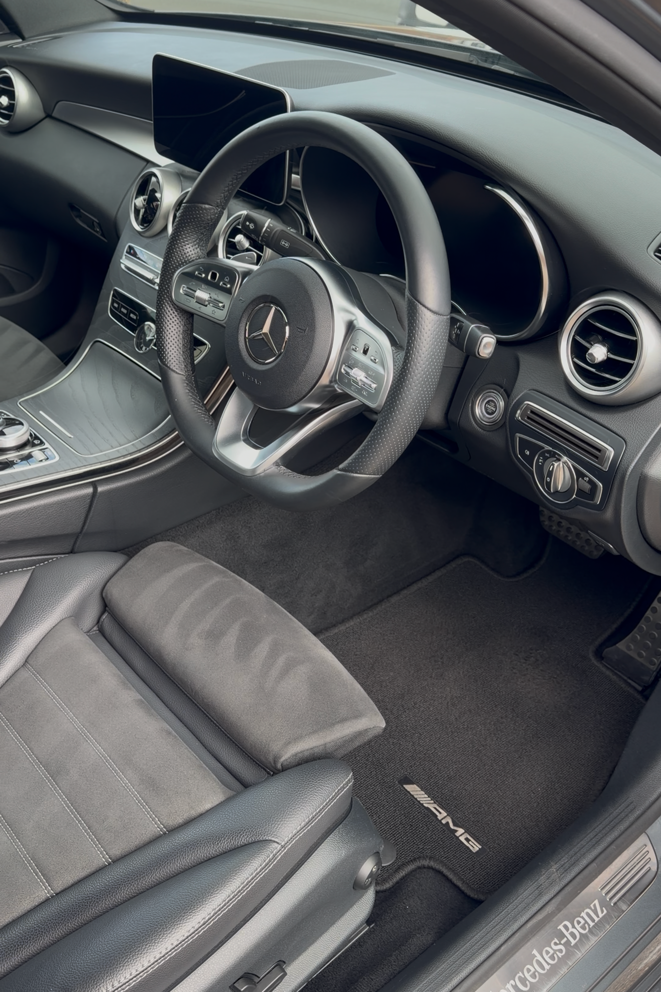 The interior of a Mercedes-Benz car, showing a black steering wheel with Mercedes-Benz logo, dashboard with digital display, central control panel, gray leather seats, and dark carpeting.