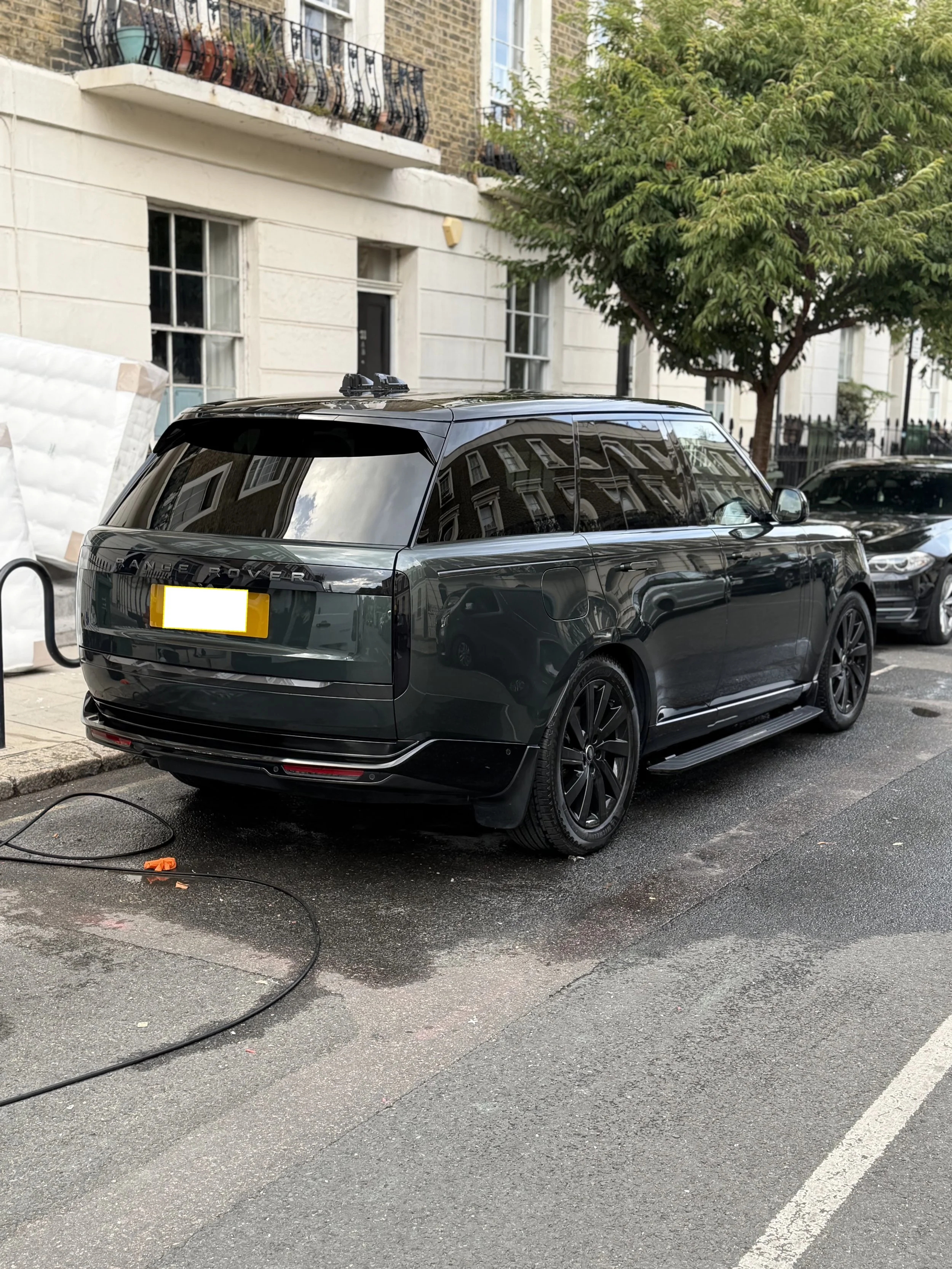 A black Range Rover parked on the street with reflections of nearby buildings on its windows, next to a building with large windows and trees, and an electric cord on the ground.