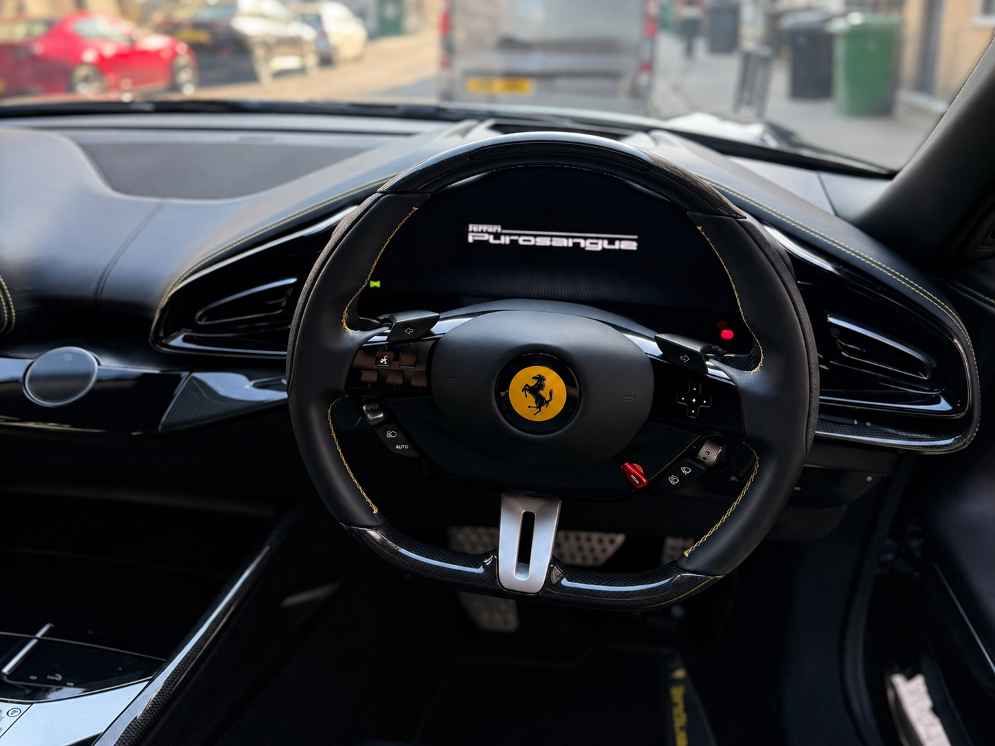 The interior of a Ferrari race car showing a steering wheel with a Ferrari logo, digital dashboard screen displaying the 'Ferrari Purosangue' logo, and dashboard air vents, with other vehicles visible outside through the windshield.