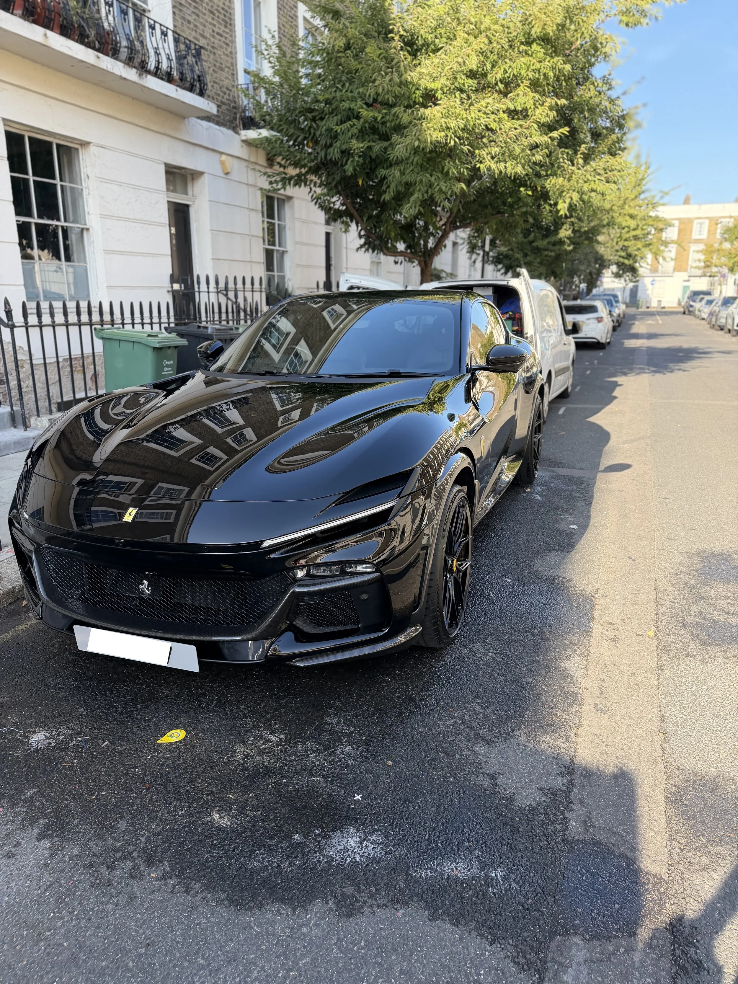 A black Ferrari sports car parked on a street with a white van behind it, residential buildings, trees, and a clear blue sky in the background.