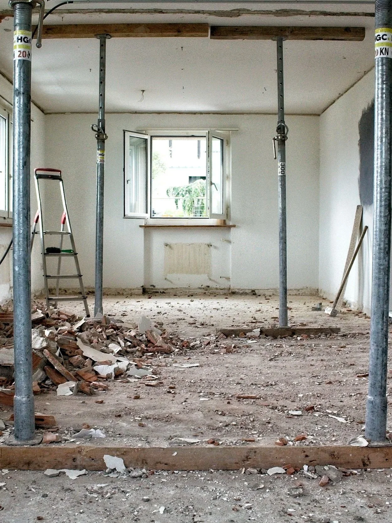 Innenraum eines renovierungsbedürftigen Zimmers mit Bauzubehör, Gerüststützen und Schutt auf dem Boden, ein Fenster ist offen, und die Wände werden repariert.