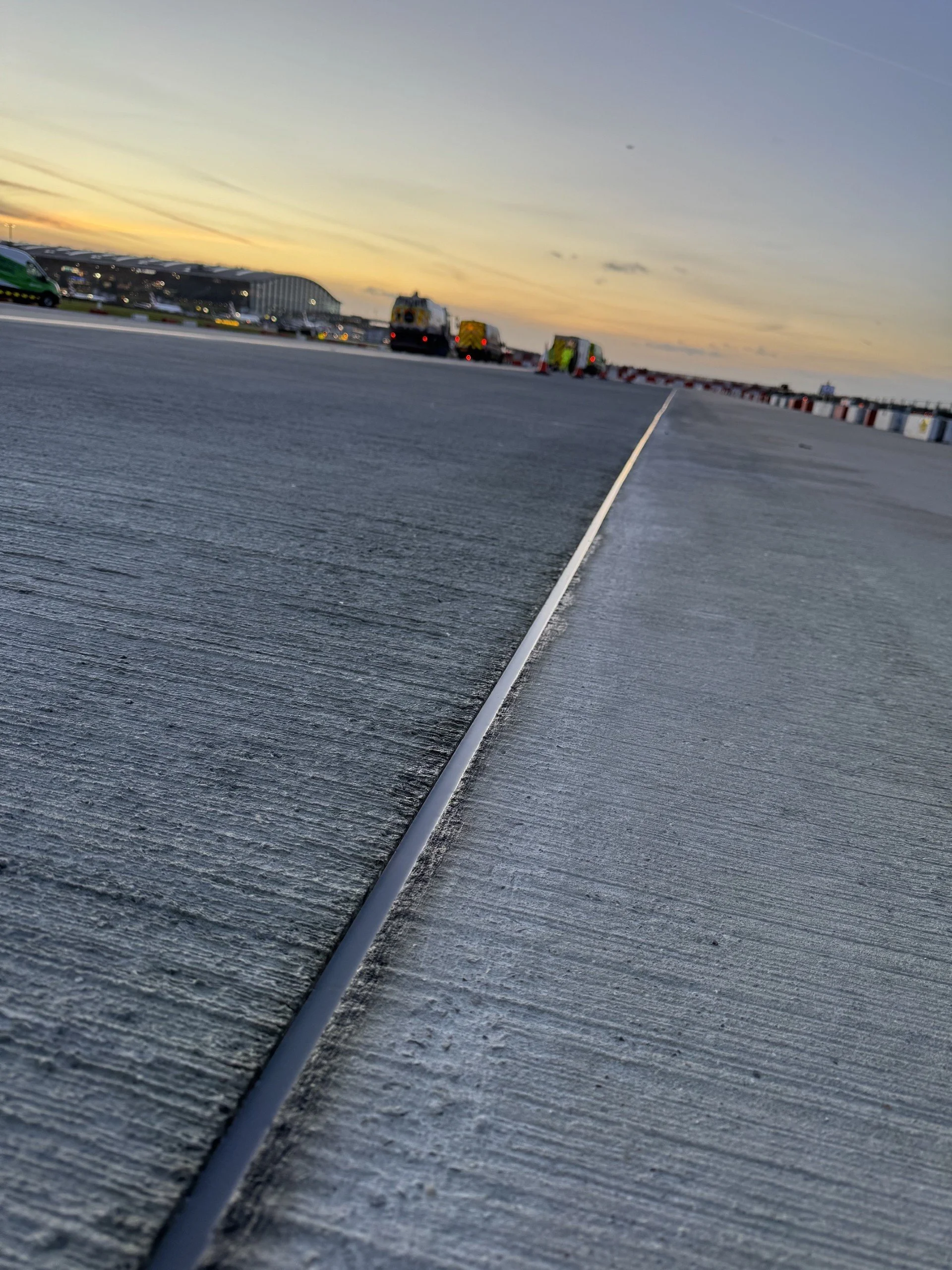 Blurry sunset view of trucks and construction equipment on a large paved surface.