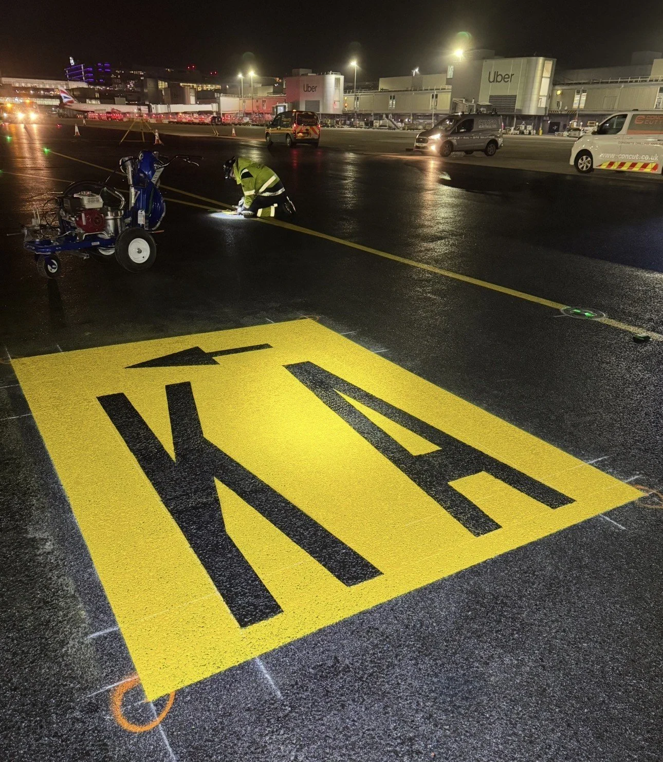 Airport runway crew member repainting a large yellow taxiway marking at night outside an Uber-branded terminal