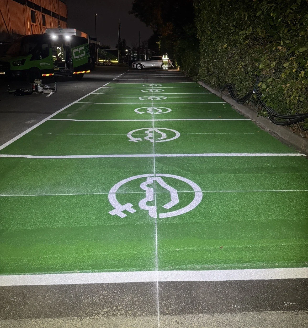 Green electric vehicle charging area with white charging symbols, service trucks, and a worker at night