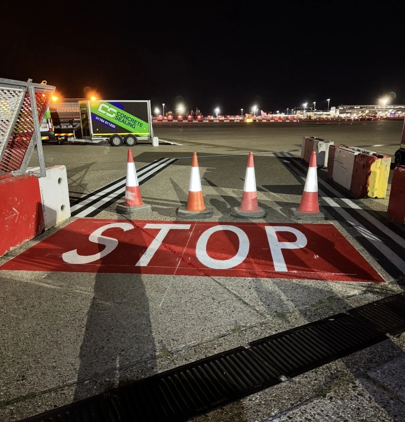 Nighttime construction area with traffic cones, red-and-white barriers, a large painted STOP marking, and a concrete sealing trailer.