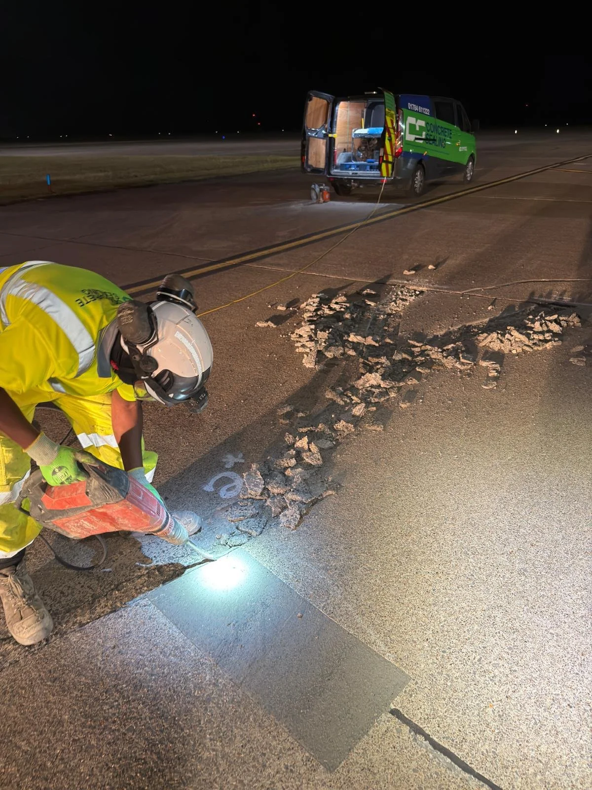 Road worker in high-visibility gear using a concrete saw to repair damaged airport runway pavement at night, with a service van nearby