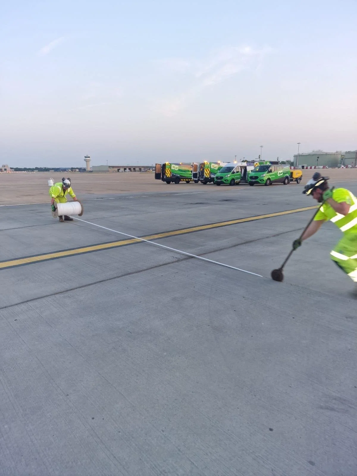 Airport ground crew in high-visibility uniforms handling equipment on the tarmac, with green service vans and an air traffic control tower in the background.