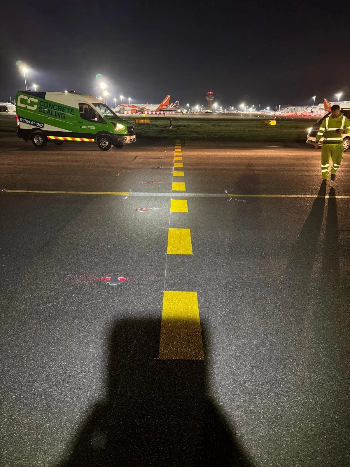 Nighttime airport apron with a green Concrete Scanning van, reflective worker, runway markings, and parked aircraft.