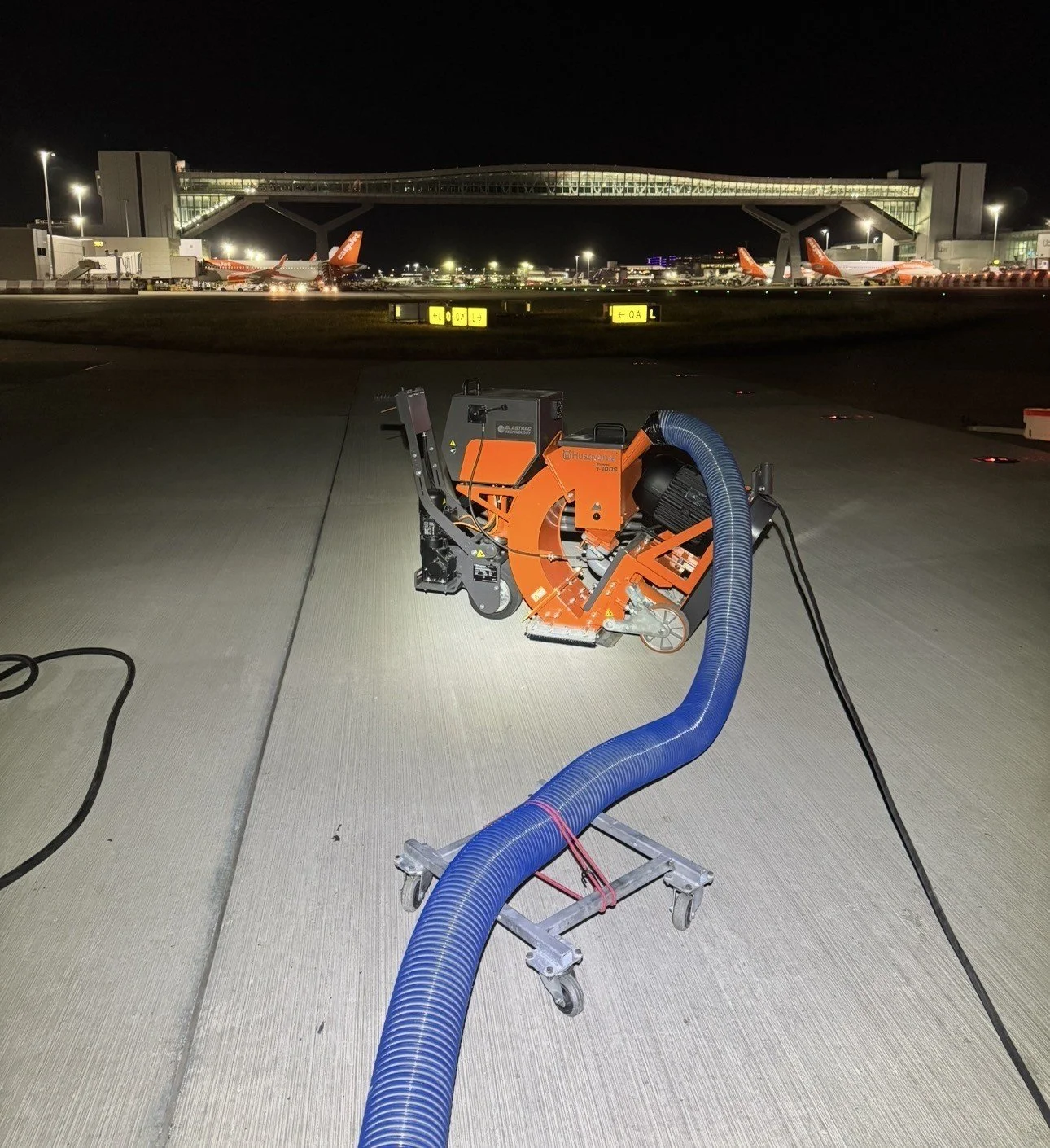 Orange Husqvarna floor grinder with a blue vacuum hose on an airport apron at night, with airplanes and a glass pedestrian bridge in the background.