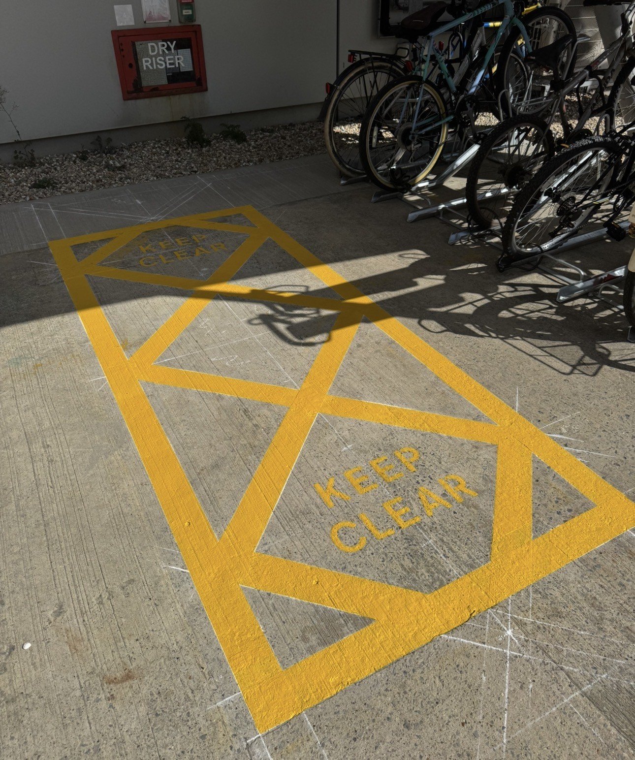 Bicycle rack beside a building with yellow crosshatched pavement marked “KEEP CLEAR” and a red “DRY RISER” cabinet.