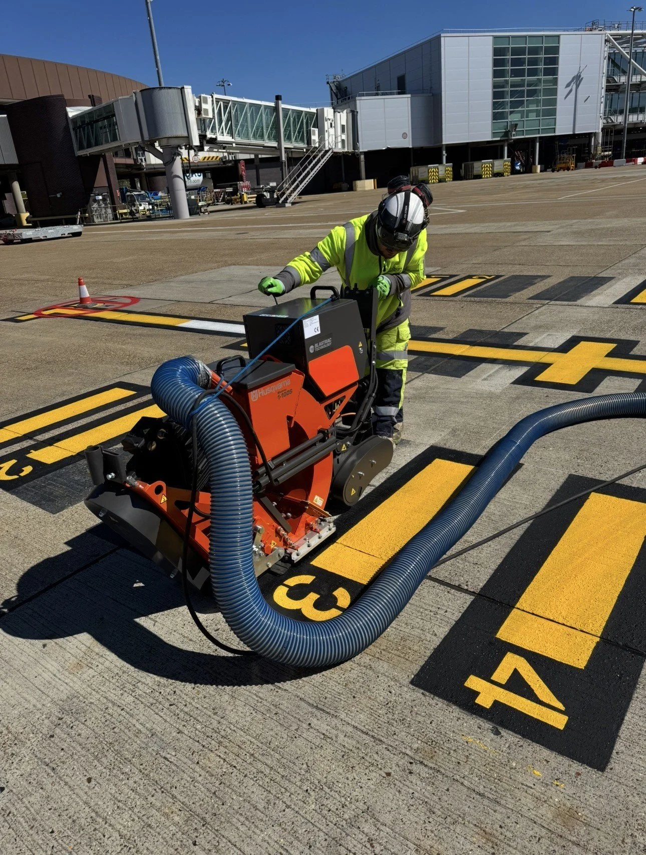 Airport ground worker operating an industrial vacuum sweeper on an airport apron beside a passenger boarding bridge