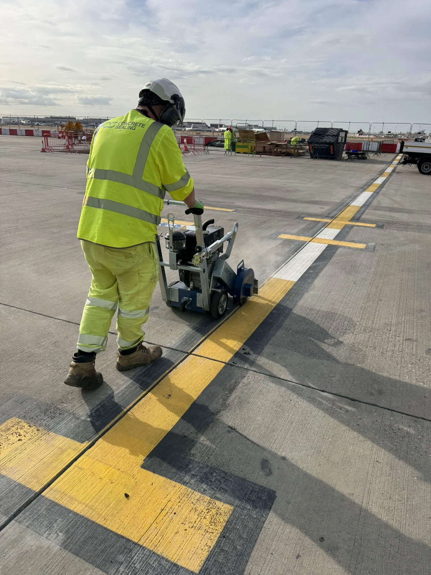 Worker in high-visibility protective gear operating concrete-cutting equipment on an airport runway.