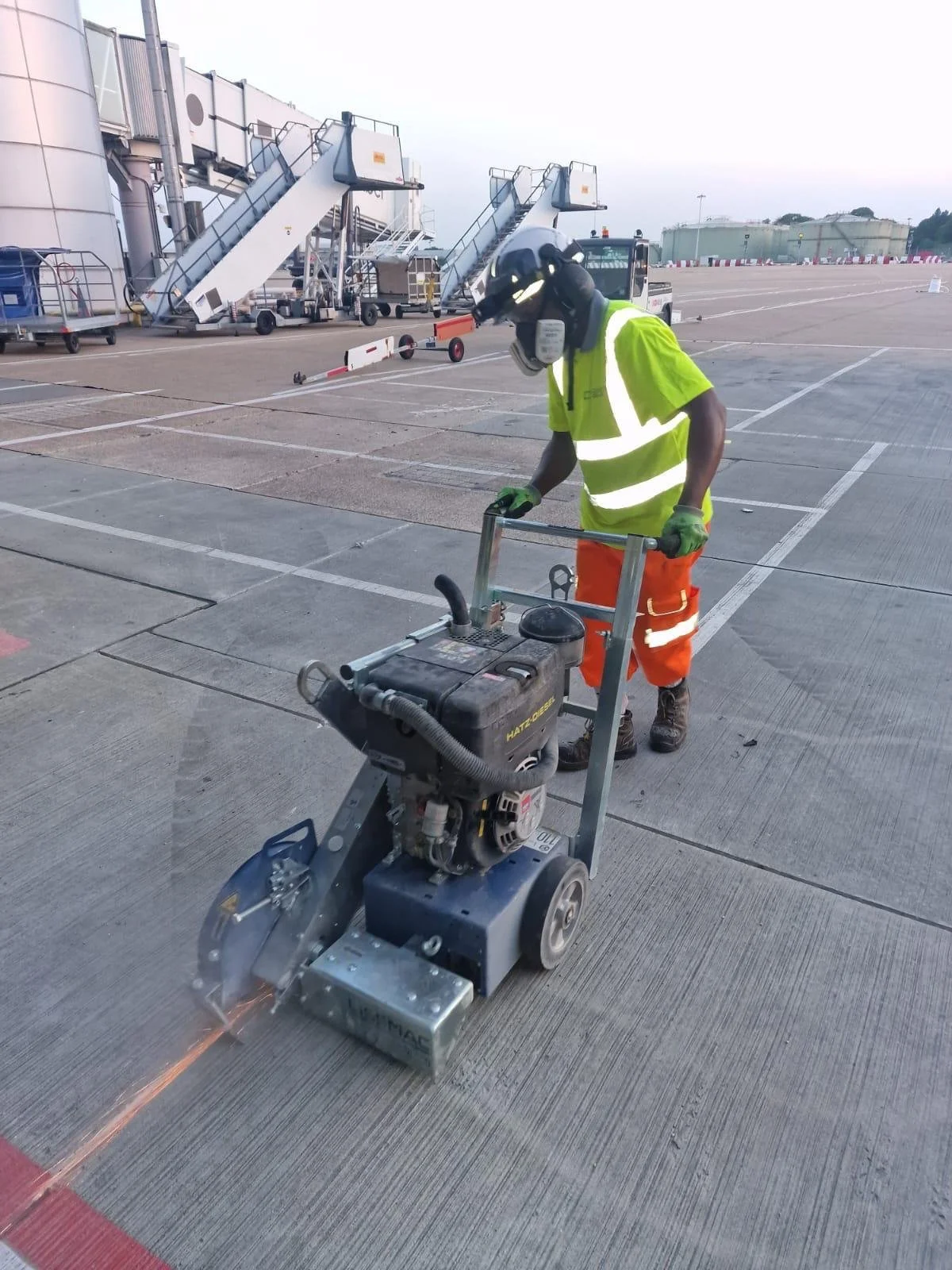 Airport worker in high-visibility clothing operating a floor-cutting machine on the airport apron near passenger boarding stairs