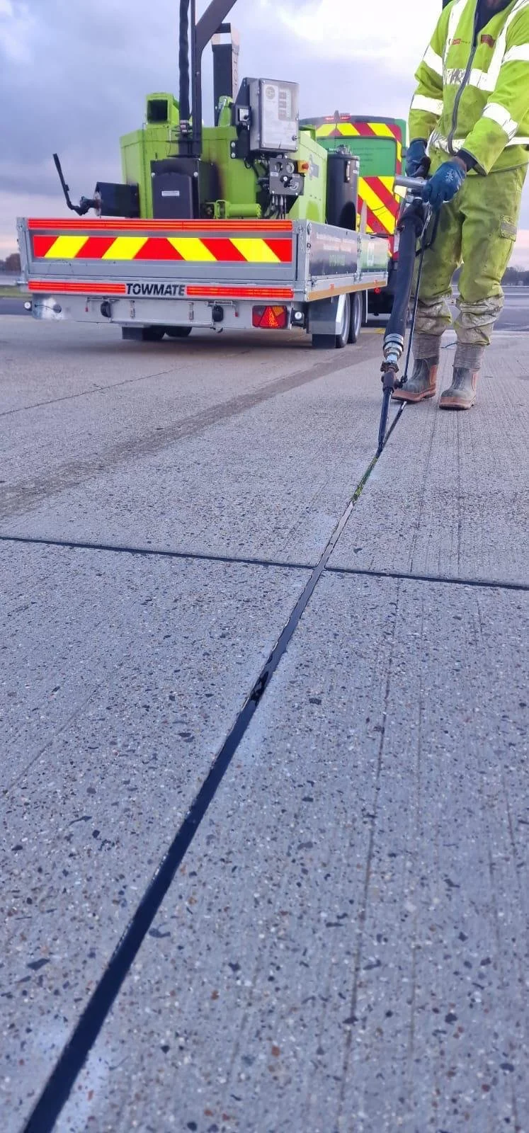Road worker in high-visibility clothing using a crack-sealing tool on concrete pavement beside a TOМMATE maintenance truck