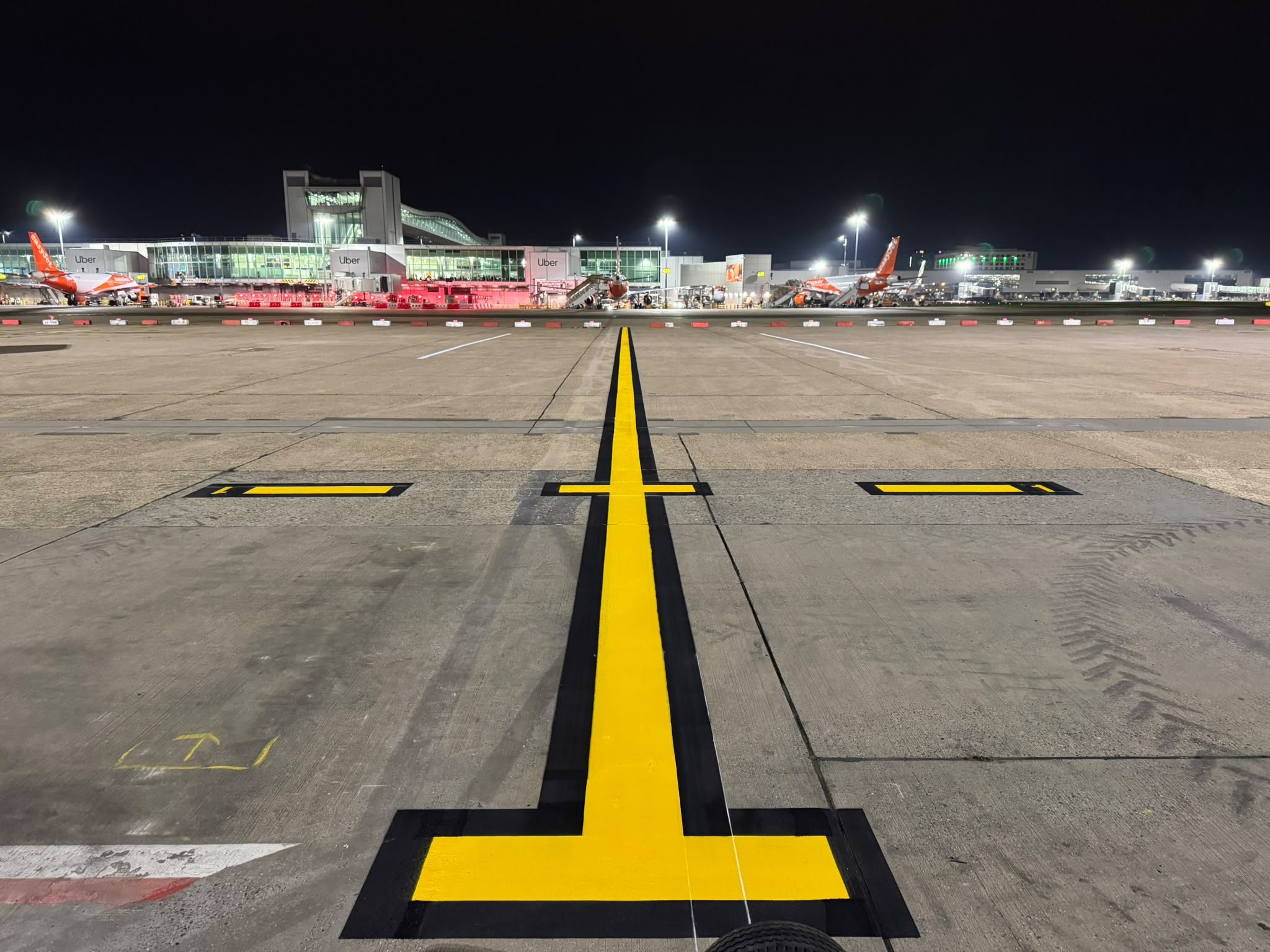 Nighttime view of an airport terminal and parked easyJet aircraft from the apron, with bright yellow taxiway markings in the foreground.