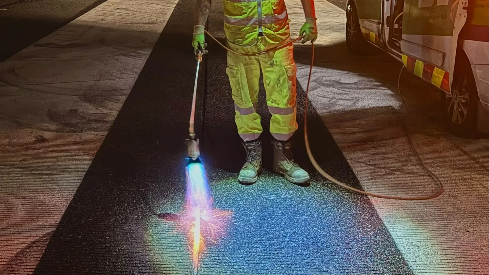 Road worker in reflective safety gear using a torch to repair asphalt beside a service vehicle