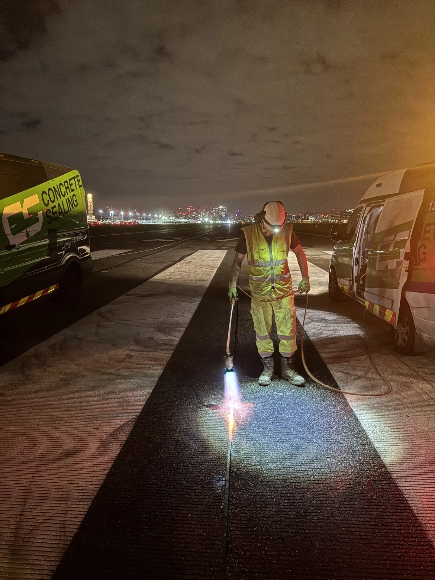 Worker in high-visibility protective gear uses a torch to seal an airport runway at night, with service vans and city lights in the background.