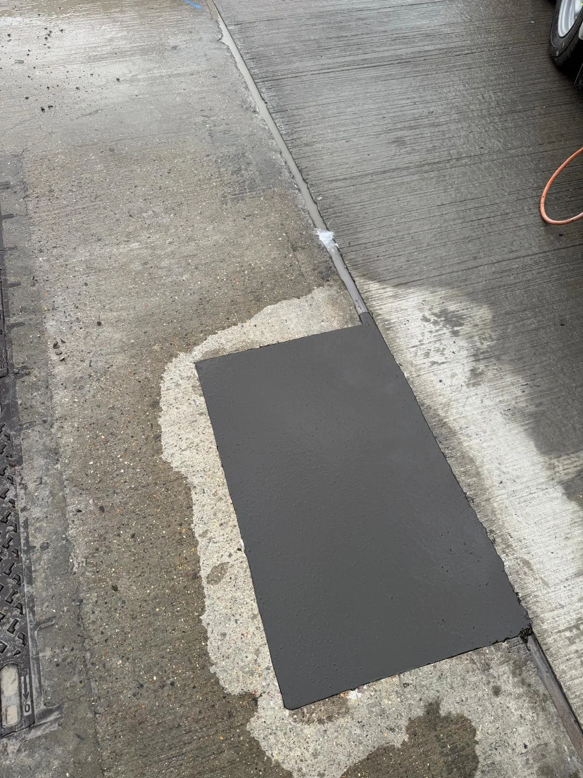 Freshly repaired rectangular section of dark concrete driveway beside a ridged concrete surface, with an orange extension cord and vehicle wheel visible.