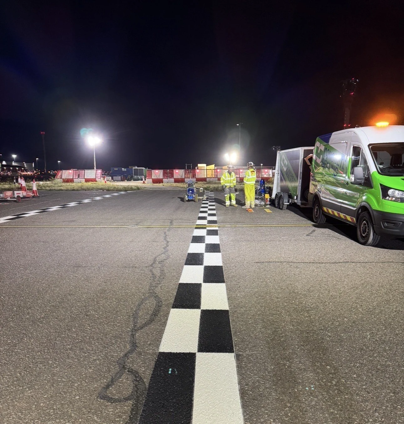 Nighttime roadwork scene with two workers in high-visibility gear, a green utility van, floodlights, traffic barriers, and a black-and-white checkered roadway marking.