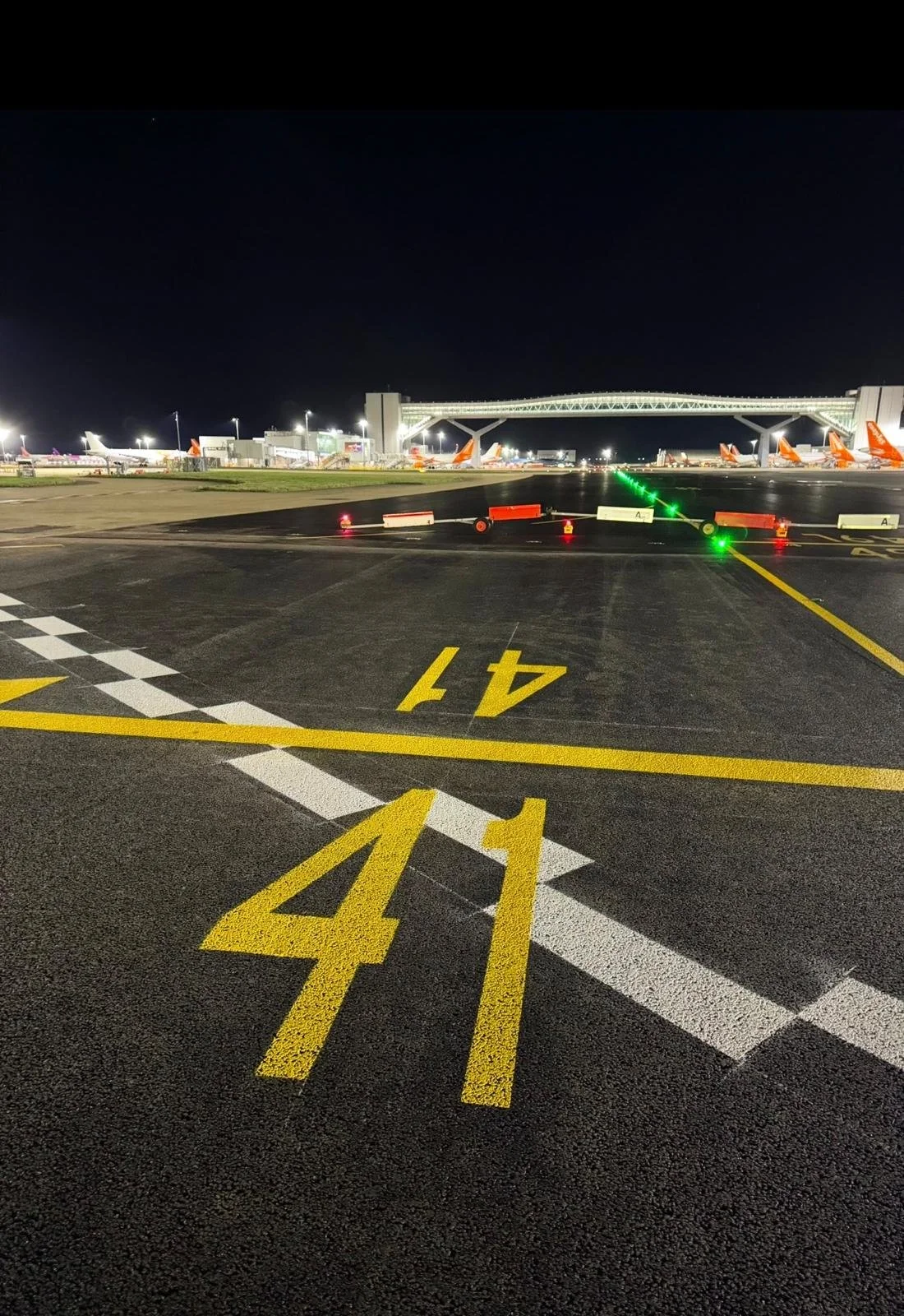 Nighttime view of an airport apron with parked commercial airplanes, illuminated terminal bridge, taxiway lights, and yellow runway markings