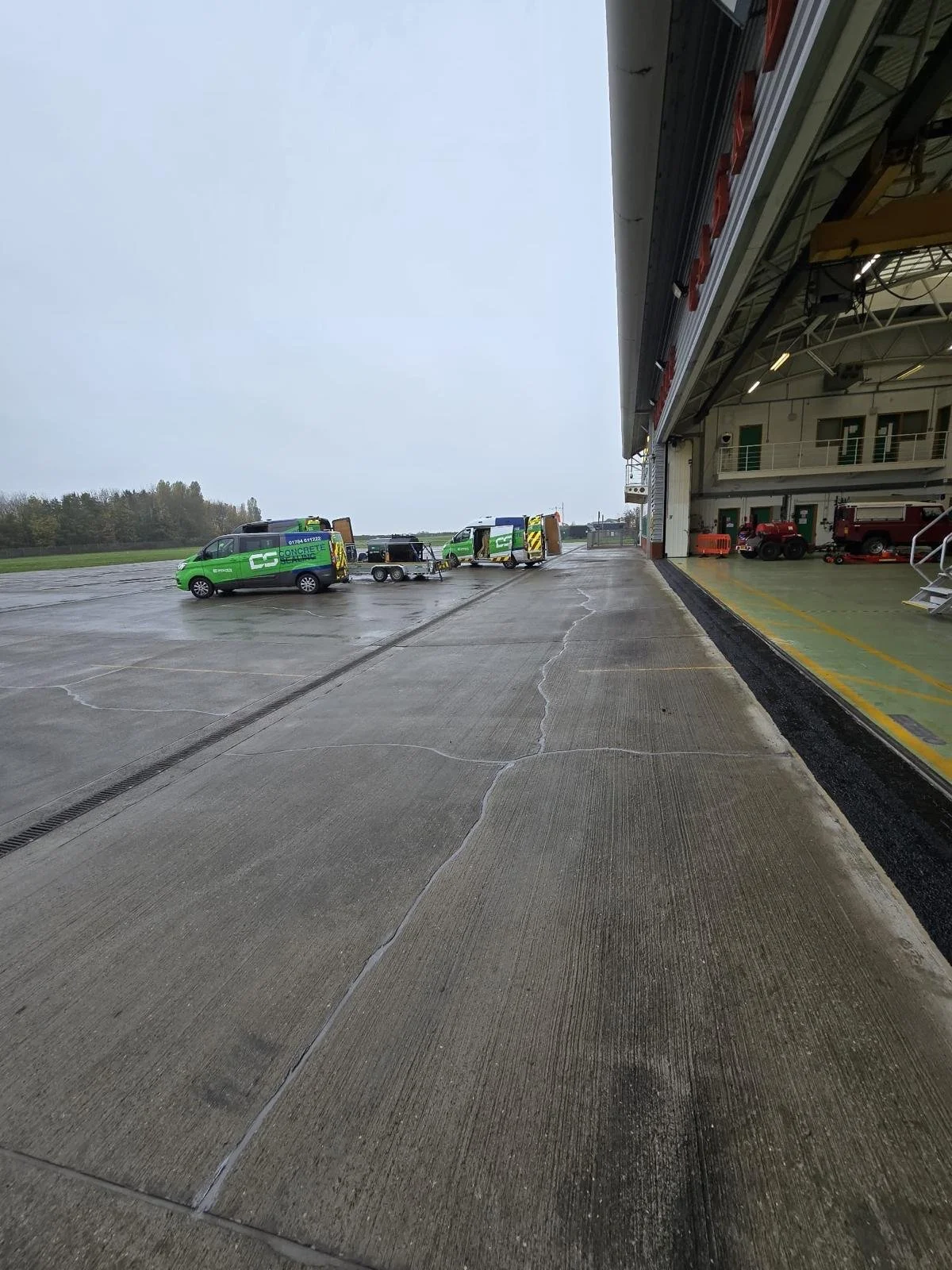 Rainy airport hangar apron with green service vans, a trailer, and fire trucks inside the open maintenance bay