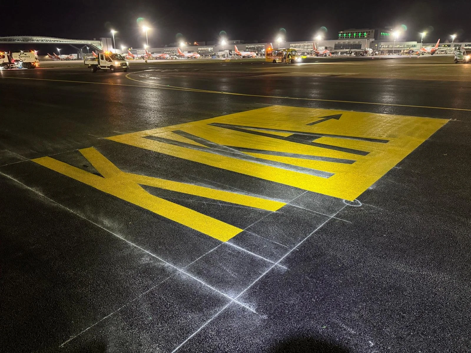 Nighttime airport apron with aircraft, ground service vehicles, terminal buildings, and bright yellow taxiway markings