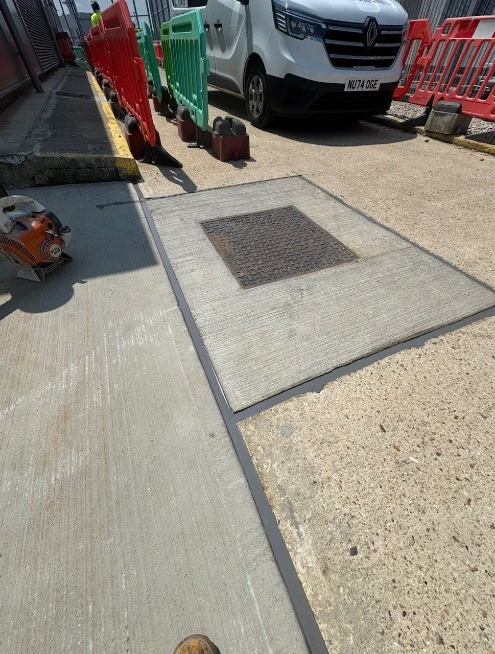 Construction site with red and green safety barriers, a white Renault van, concrete paving slabs, and a utility access cover