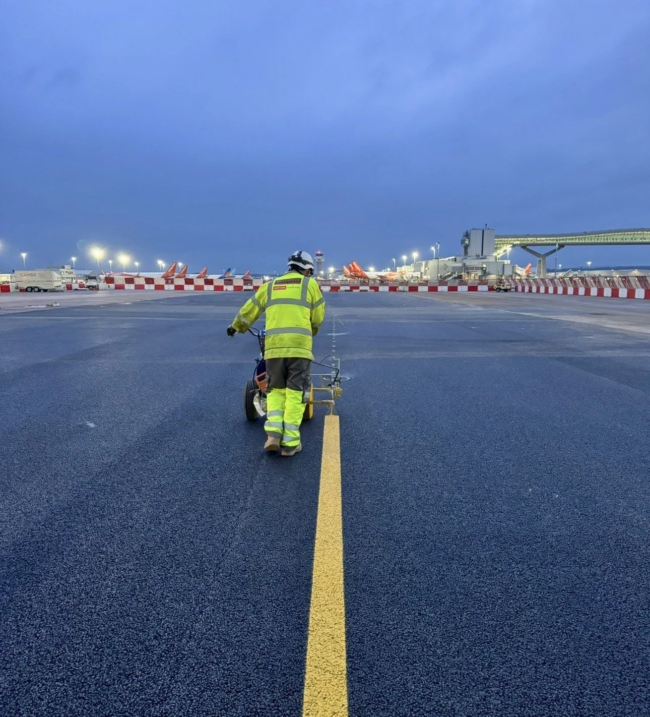 Airport ground worker in high-visibility gear pushes equipment along a runway at dusk, with parked aircraft and terminal buildings in the background.