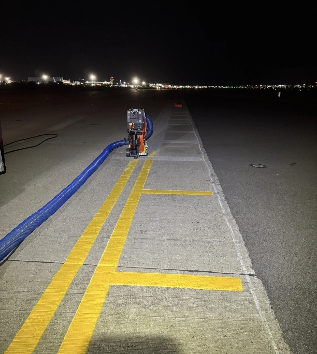 Airport runway at night with ground service equipment, a large blue hose, yellow pavement markings, and illuminated terminal buildings in the background.