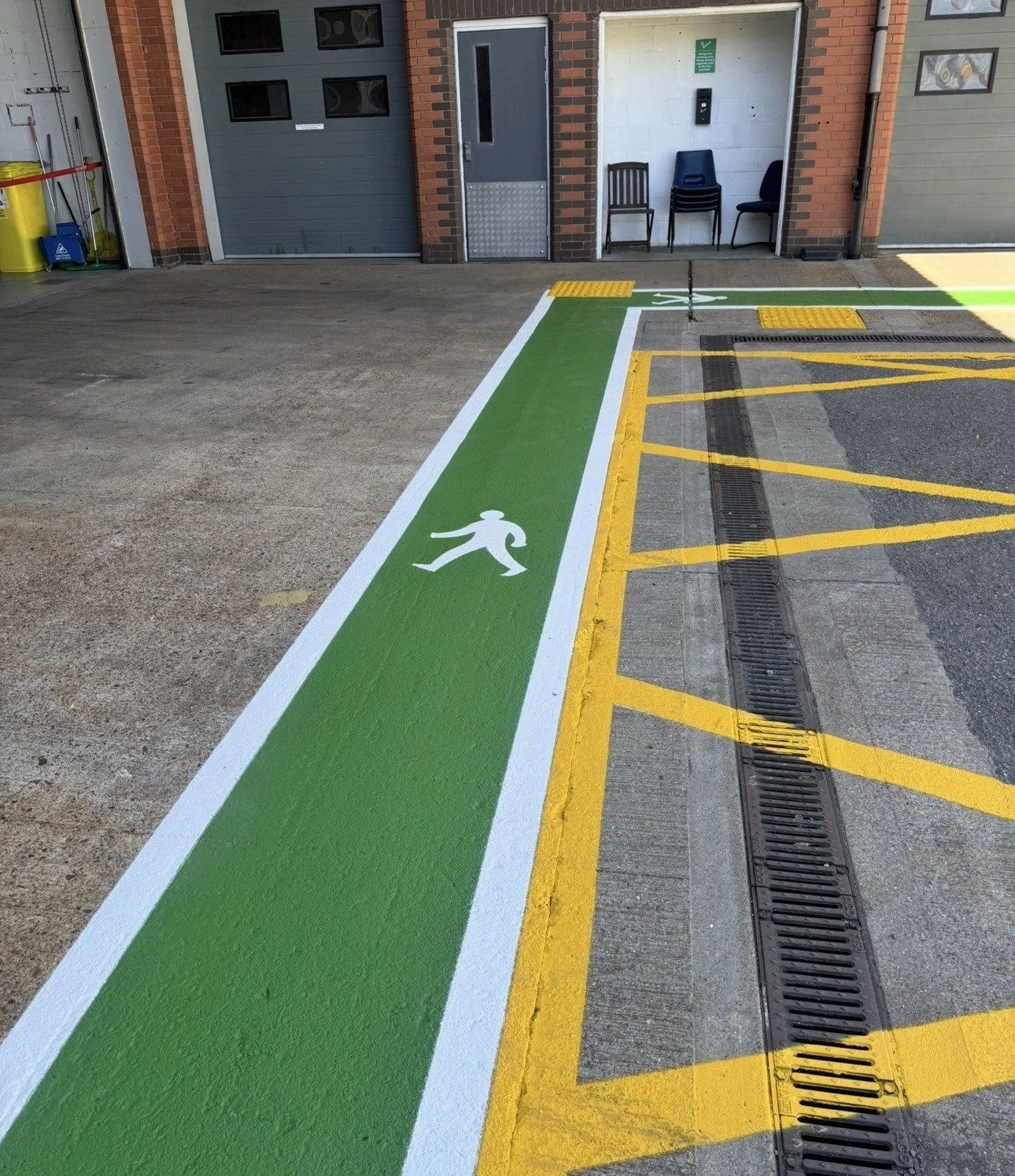 Green pedestrian walkway with white walking figure, yellow striped pavement, tactile crossings, and building entrances