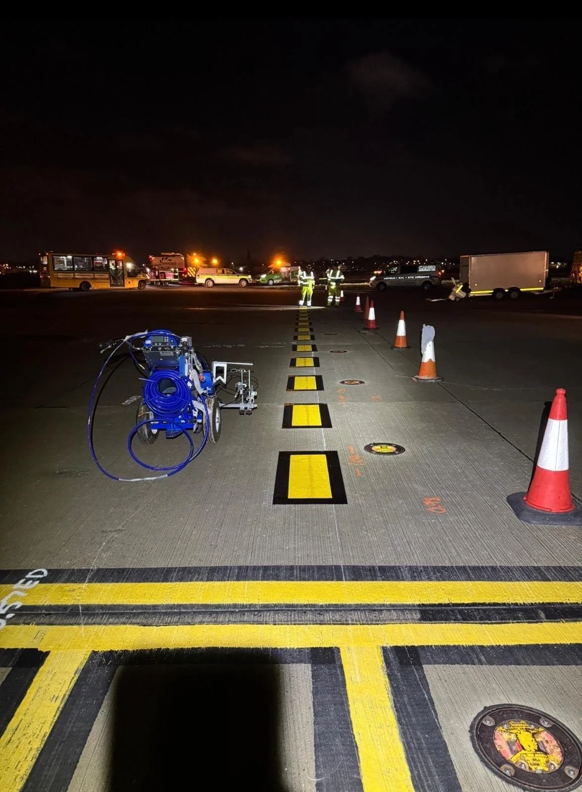 Nighttime airport runway maintenance with workers, emergency vehicles, traffic cones, and blue ground equipment.
