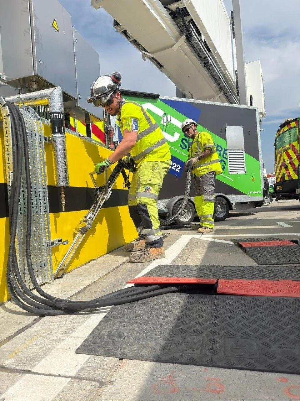 Two workers in high-visibility clothing operate industrial equipment beside a large service vehicle.