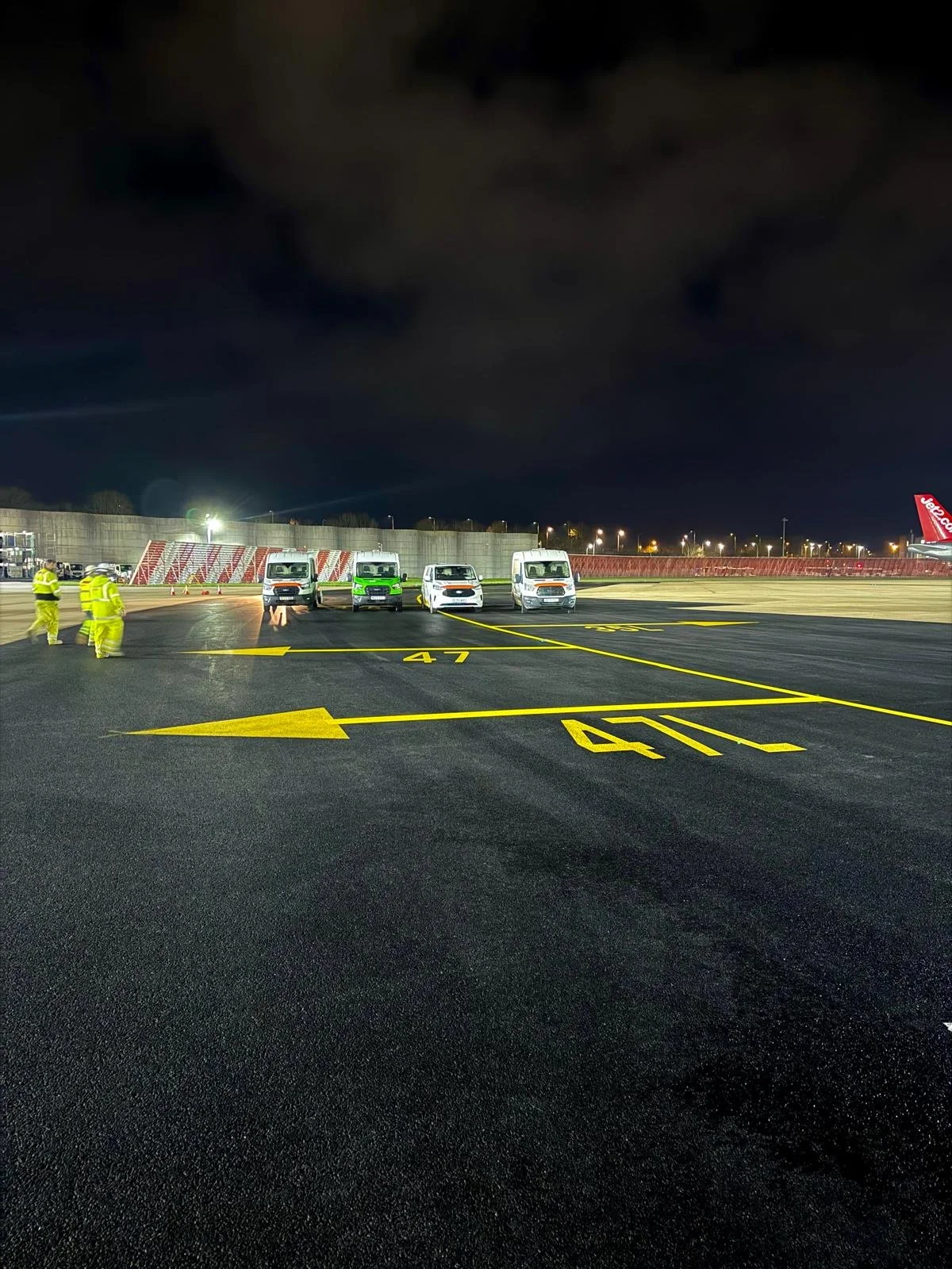 Airport tarmac at night with service trucks, ground crew in high-visibility uniforms, and yellow directional markings.
