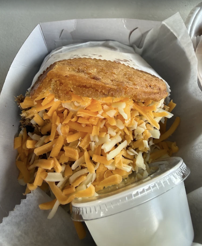 A fish sandwich topped with shredded cheese, served with a side of creamy sauce in a plastic cup.