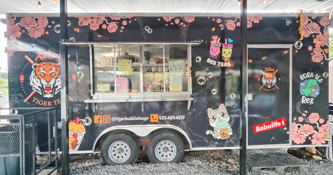 A black food truck decorated with pink flowers, cartoon animals, and colorful drink illustrations. The truck advertises bubble teas with contact info and social media handles.