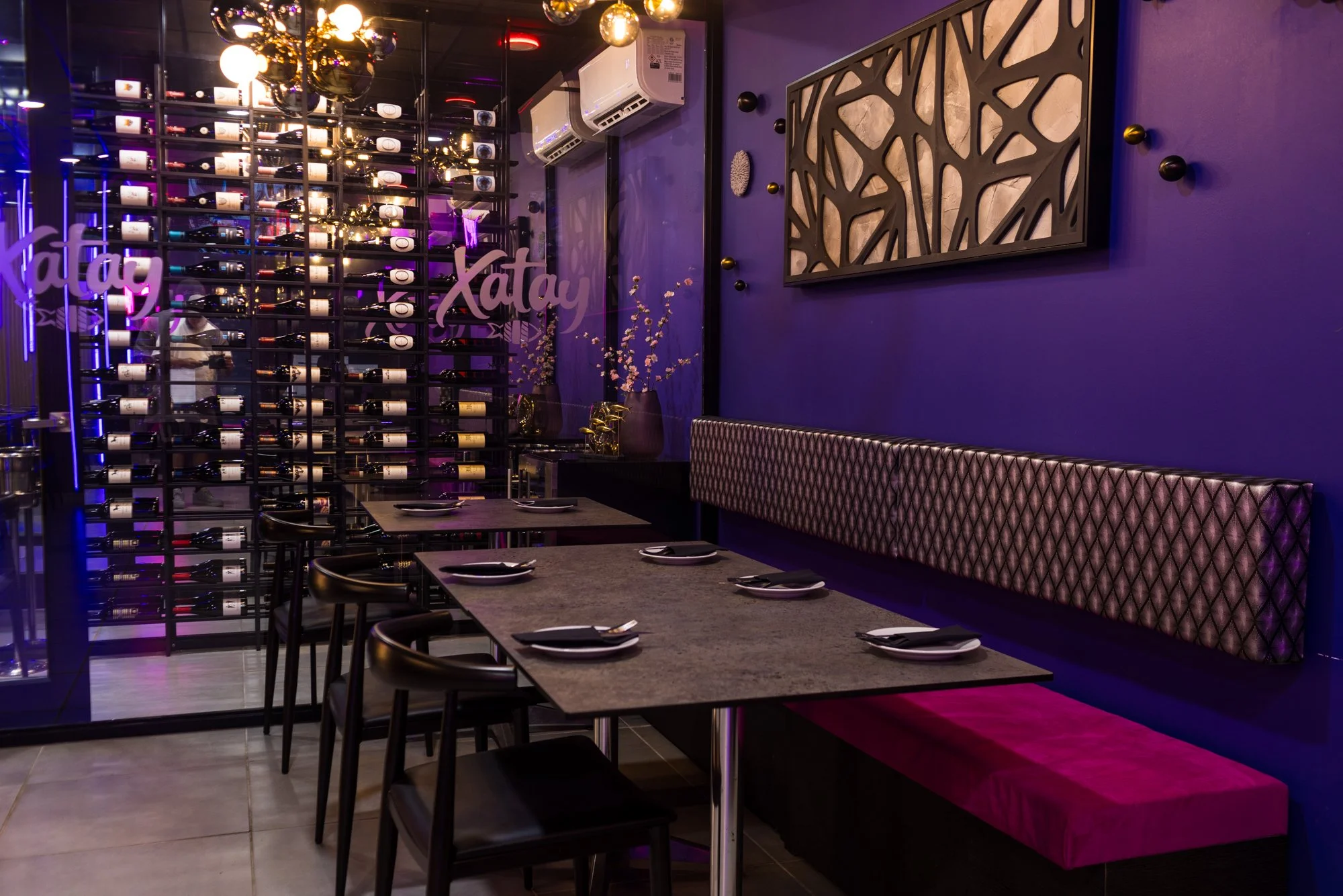 Interior of a modern restaurant with purple walls, a decorative wall hanging, a wine display wall, and a table set with plates and utensils.