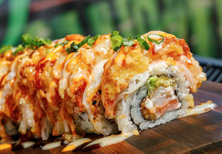 Close-up of a sushi roll topped with orange sauce, sesame seeds, chopped green onions, and drizzled with a creamy sauce on a wooden board.