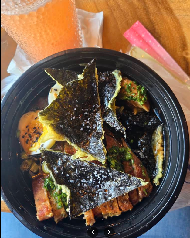 A black bowl of Japanese ramen noodles topped with seaweed, an egg, sliced pork, green onions, and other ingredients. There is a glass of iced tea and a pink spoon in the background.