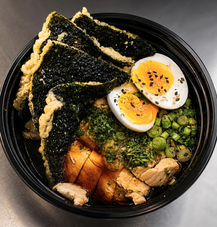Soup bowl with roasted chicken, soft-boiled eggs sprinkled with black sesame seeds, green onions, seaweed sheets, and chopped herbs.
