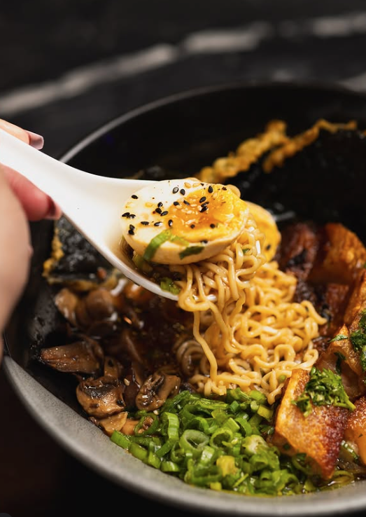 Spoon lifting cooked ramen noodles with chopped green onions, sliced mushrooms, soft-boiled egg, and pieces of pork belly in a black bowl.