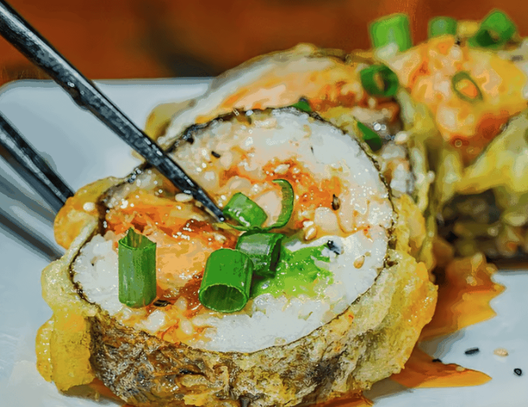 Close-up of a sushi roll with white rice, orange sauce, green onions, and fried tempura batter, served on a white plate with chopsticks.