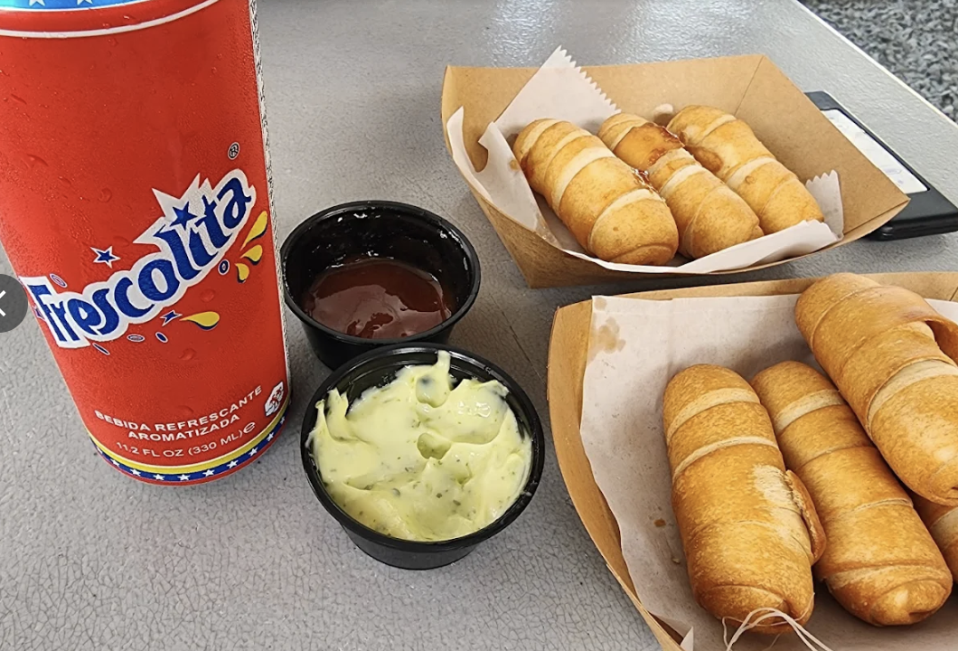 A tray of six taquitos, a basket of nine taquitos, a small container of guacamole, a small container of red sauce, and a can of Frescolita soda on a gray table.