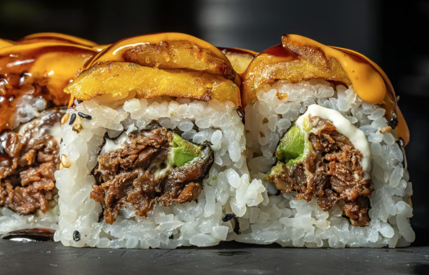 Close-up of two sushi rolls with cooked shredded beef, avocado, topped with sliced fried plantains, drizzled with caramel and spicy mayo sauces.