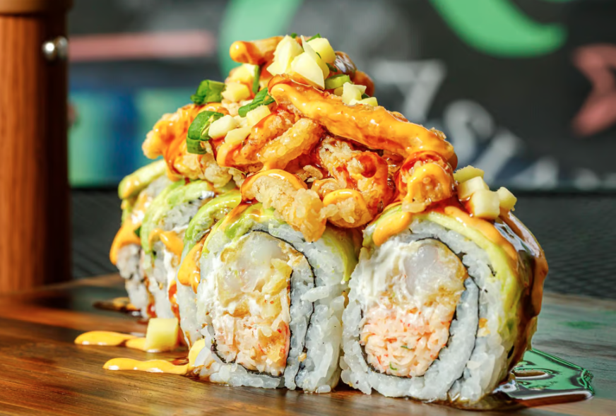 Close-up of a sushi roll topped with fried fish, chopped vegetables, and sauces, served on a wooden board.