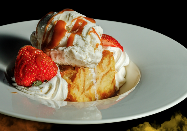 A dessert plate with a slice of bread pudding topped with a scoop of vanilla ice cream drizzled with caramel sauce, garnished with fresh strawberries and whipped cream.