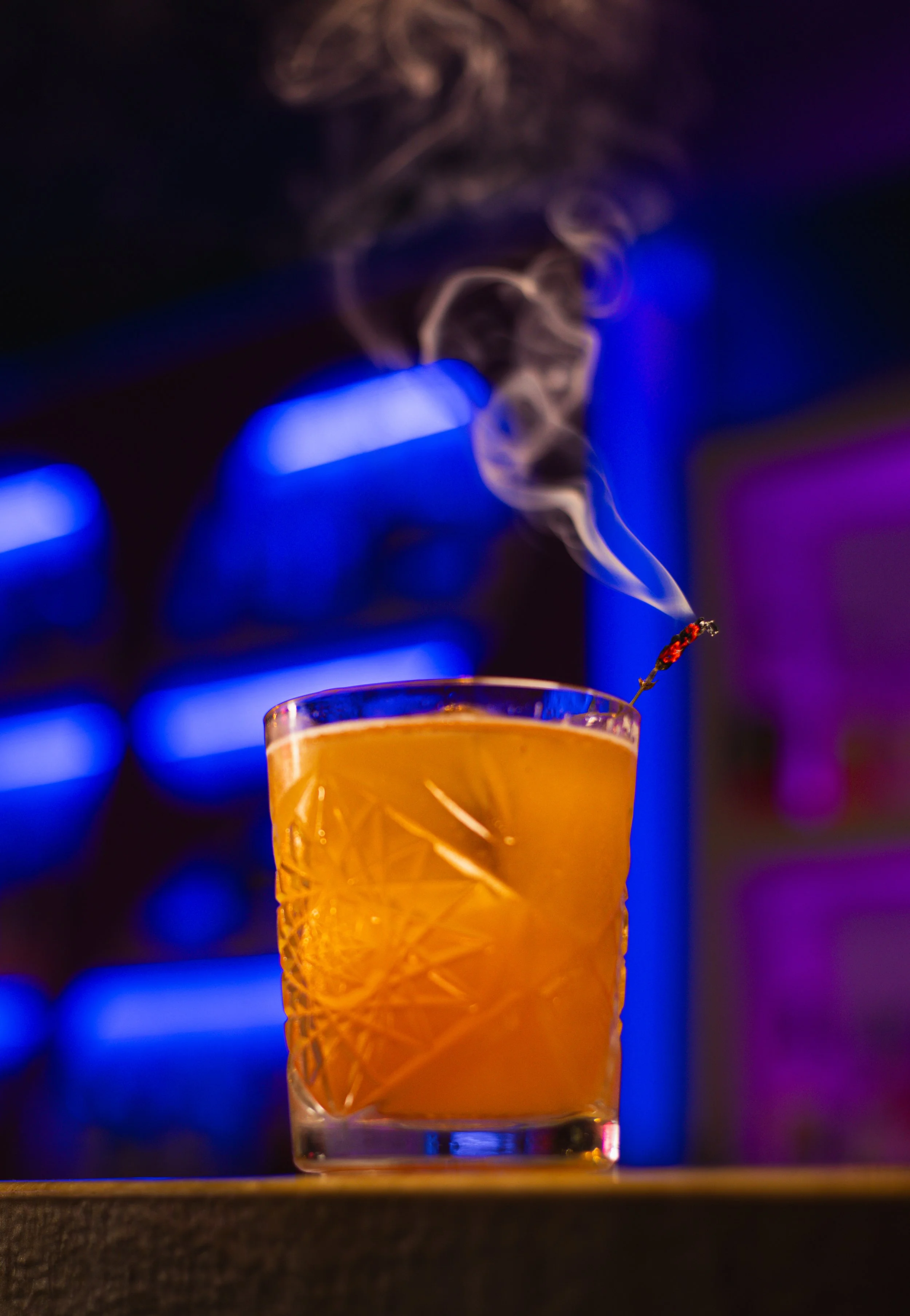 A glass of orange-colored cocktail with ice and a lit cigarette above it, emitting smoke, against a background with blue and purple lights.