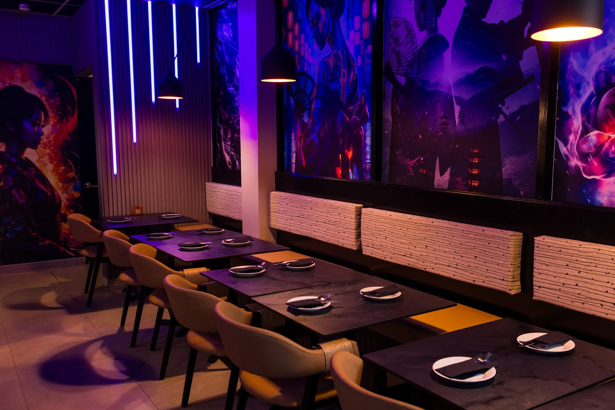 A restaurant interior with tables set for dining, featuring multiple chairs, a long booth with cushions, dark tabletops with plates and utensils, and vibrant wall art illuminated by purple and blue lighting, including neon lights and anime-style mura