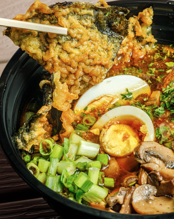 Close-up of a bowl of ramen with sliced boiled eggs, chopped green onions, sliced mushrooms, and a piece of fried fish with batter and chopsticks.