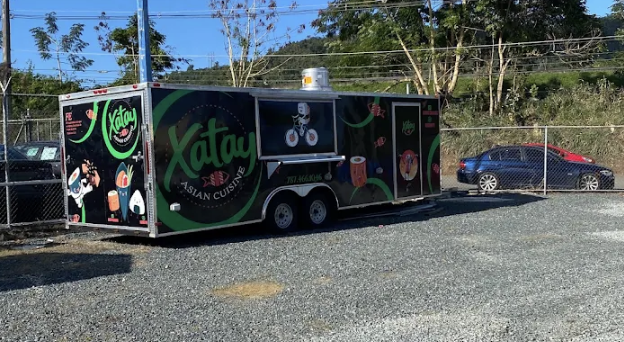 A black food truck with green and white branding for Xatay Asian Cuisine parked on gravel, with a chain-link fence and trees in the background, and two cars parked nearby.